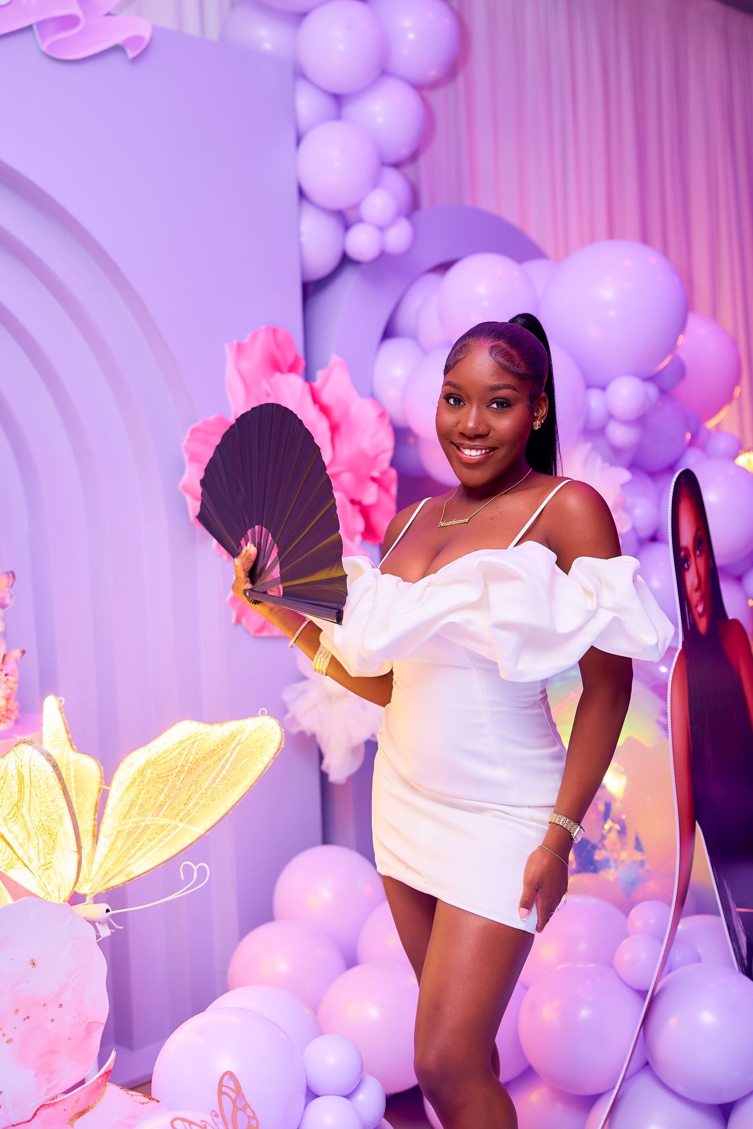 Young woman in white off-shoulder dress holding a black fan at a purple balloon-decorated party.
