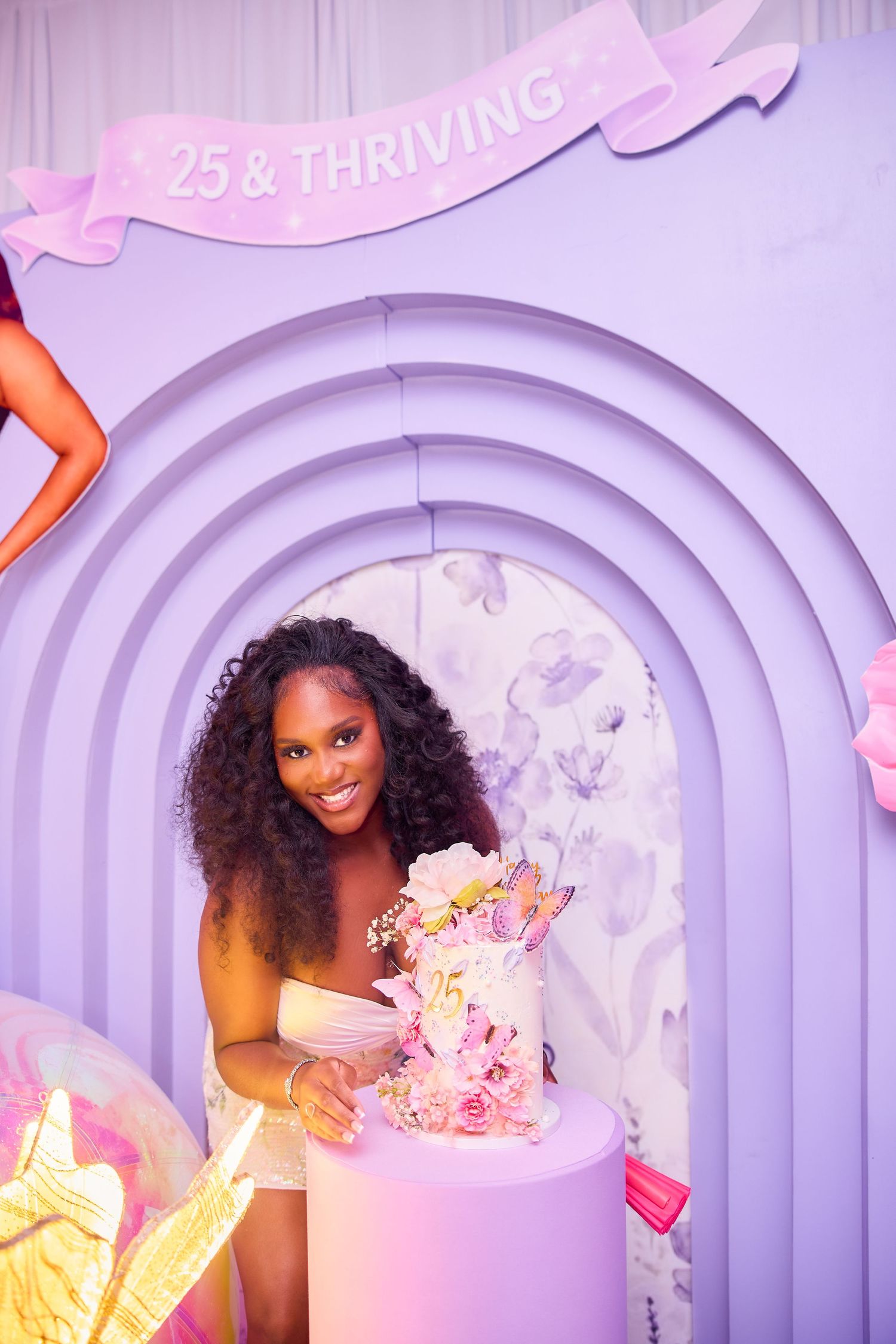 Woman smiling at a 25 & Thriving birthday party with a floral cake on a pink pedestal against a purple arch backdrop.