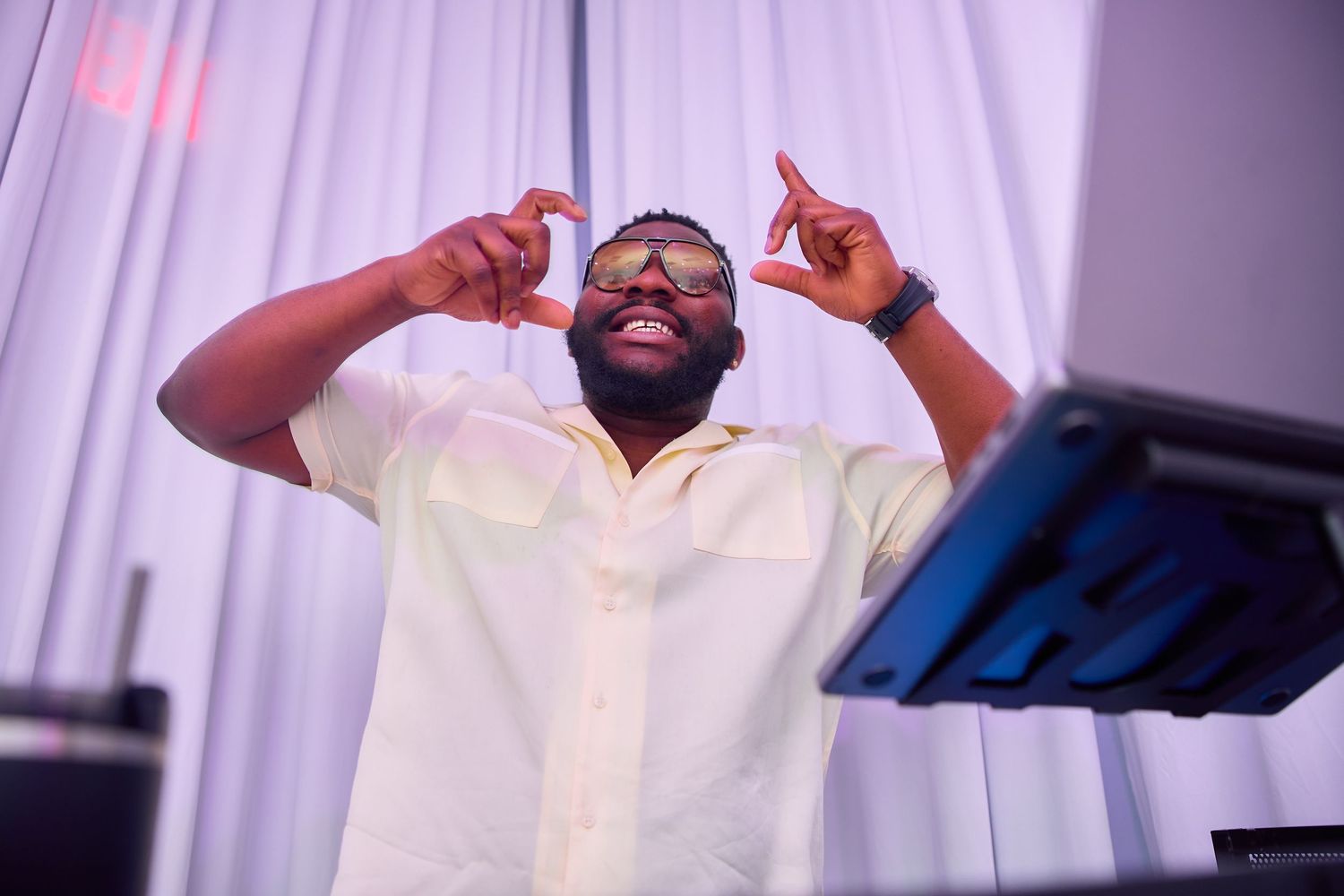 Man in yellow shirt and gold sunglasses posing at a DJ booth at a nighttime event.