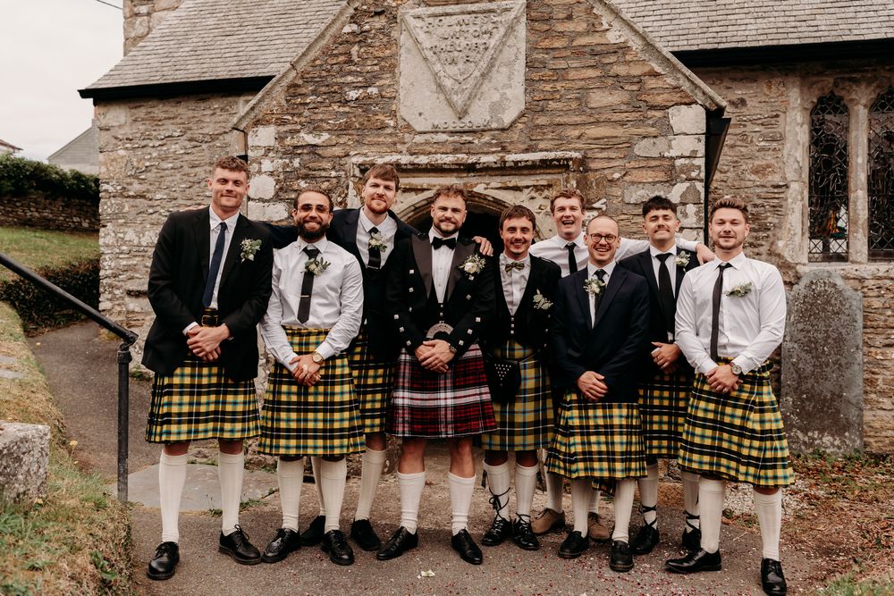 Wedding party at rustic stone venue wearing mix of traditional kilts and beige suits for formal group photos.