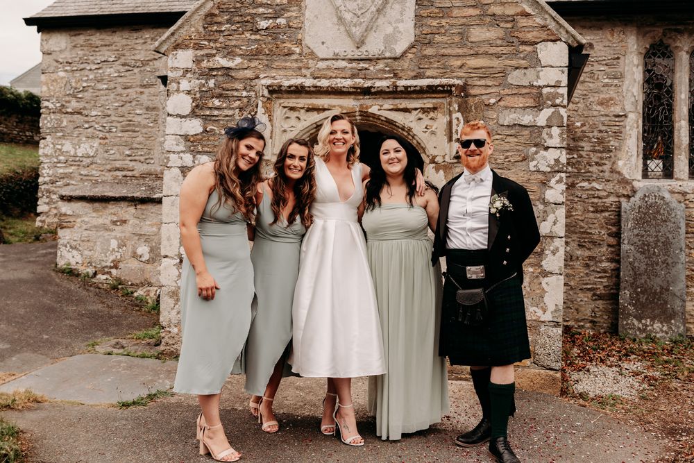 Wedding party members in sage green dresses and suits pose together at a rustic stone church entrance.