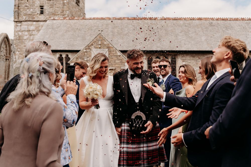 A crowd gathers for a wedding celebration outside a historic stone church in natural lighting.