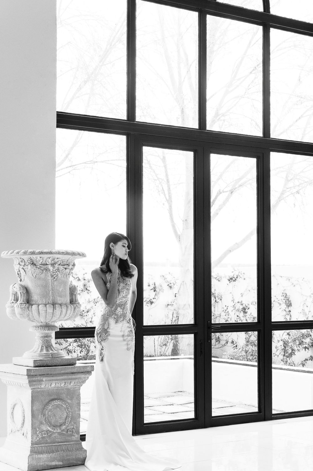 A person in an elegant white wedding dress stands in a bright modern room with large windows and decorative columns.