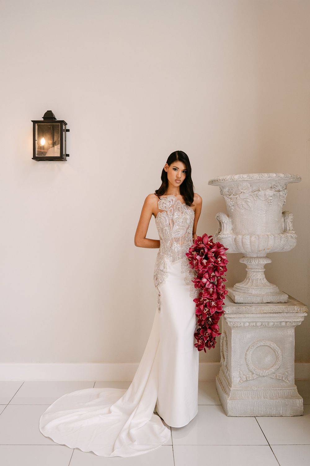 A bride in a white gown holding a cascading burgundy orchid bouquet poses against a light colored wall with decorative urns.
