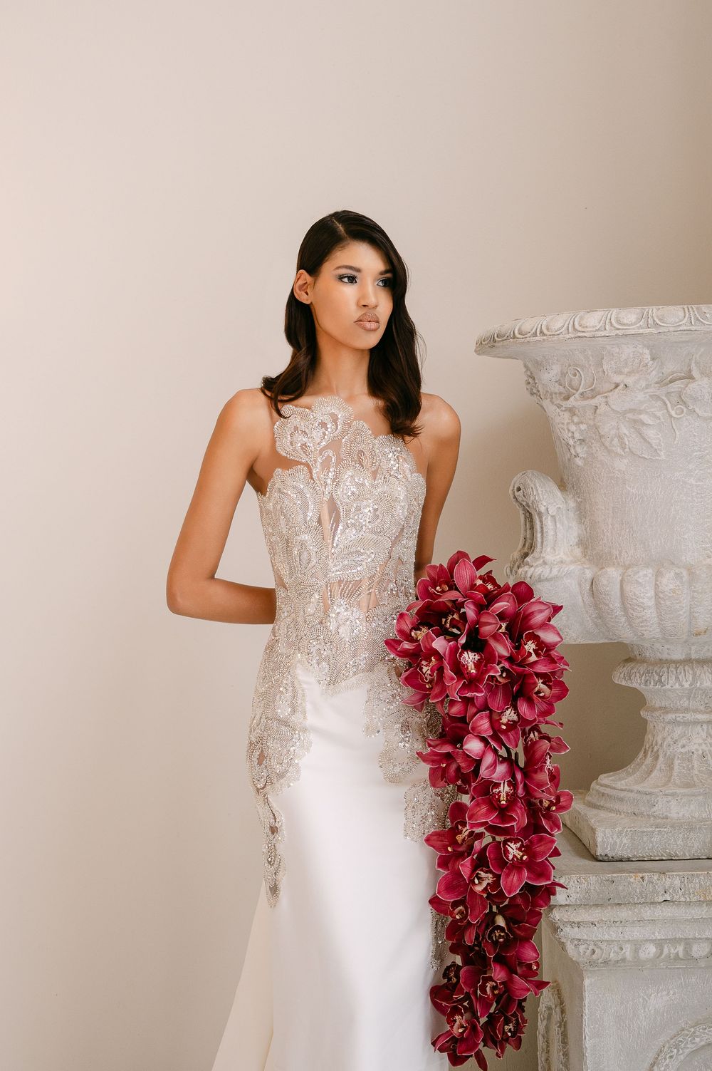 Elegant bride in white lace gown carries cascading burgundy orchid bouquet against neutral backdrop with ornate urns.