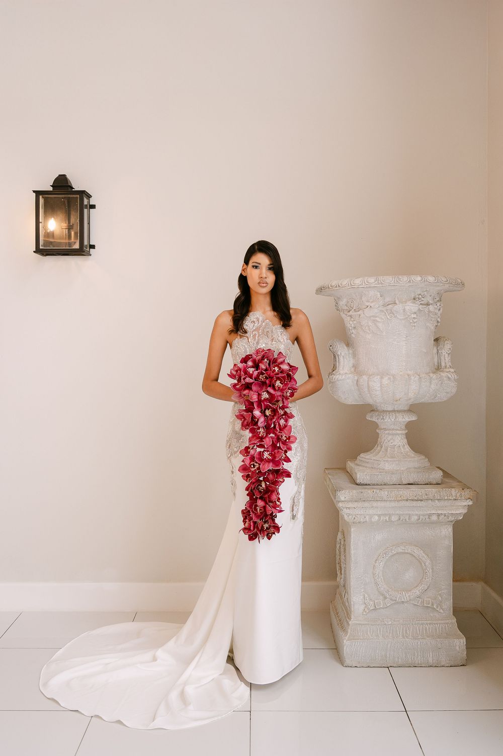 Elegant bride in flowing white gown holds cascading burgundy orchid bouquet against neutral studio backdrop with decorative urn.