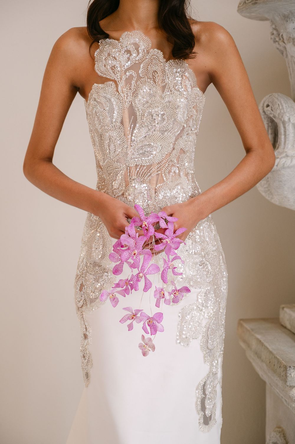 A strapless white wedding dress with intricate lace detailing and pink orchid flowers held at the waist.