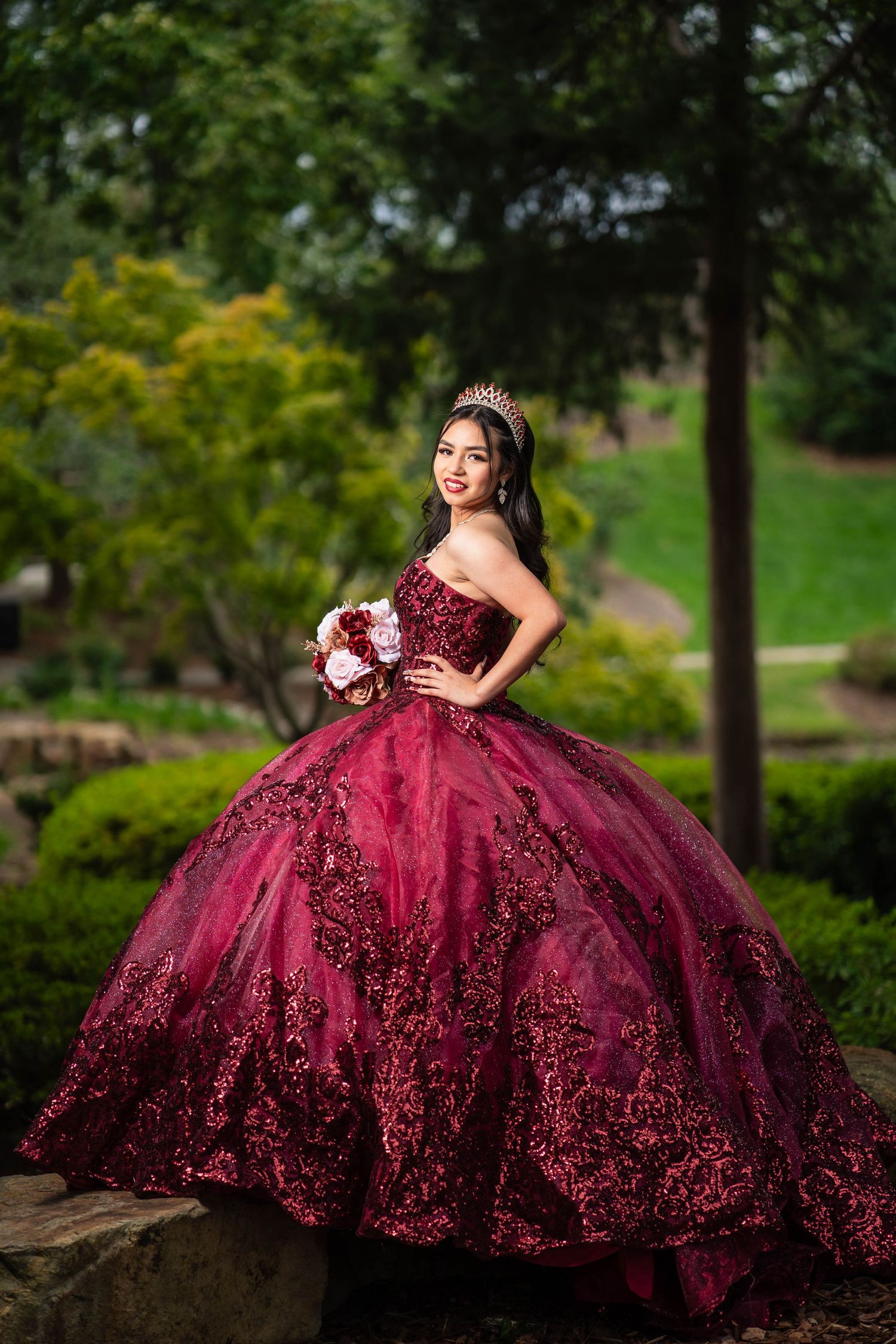 A burgundy quinceañera ball gown with intricate beading sparkles in an outdoor garden photoshoot.