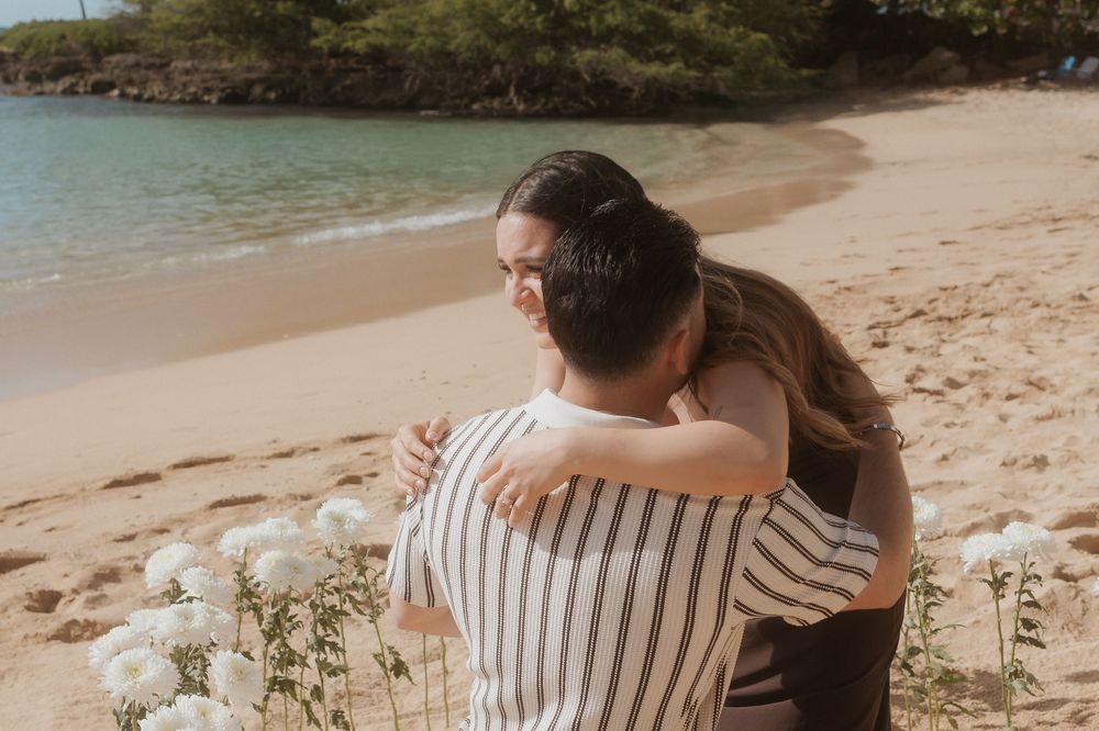 A couple sitting on a sandy beach shore wearing casual attire with the ocean waves in the background.