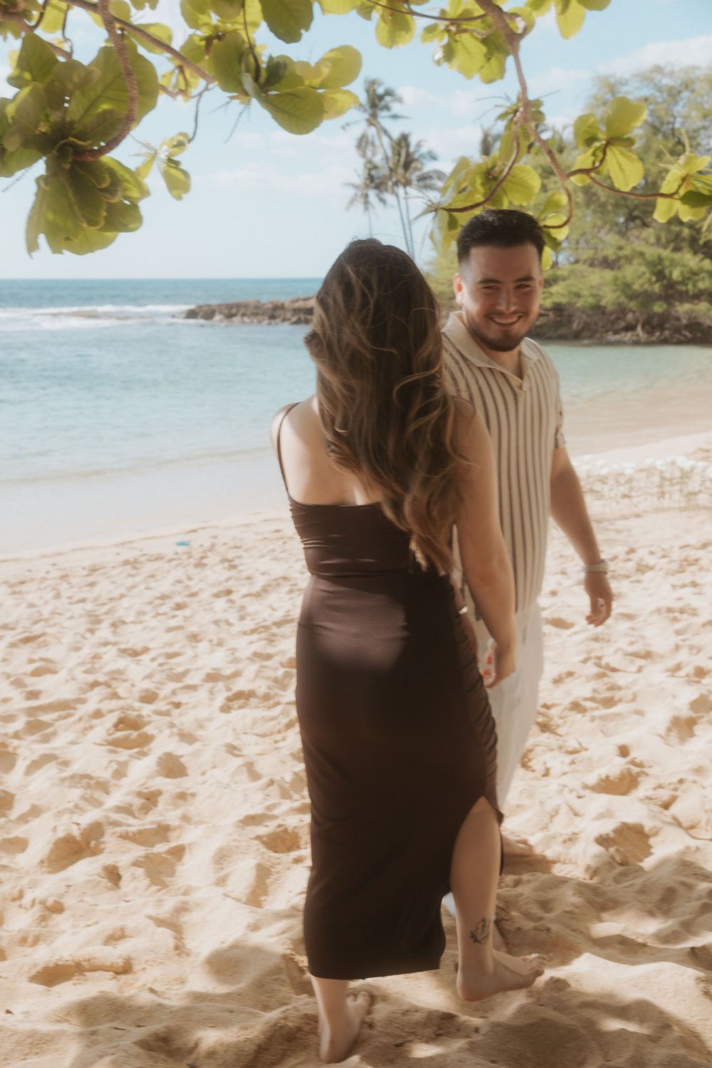 Couple walking hand in hand along a tropical beach with white sand and palm trees in the background.