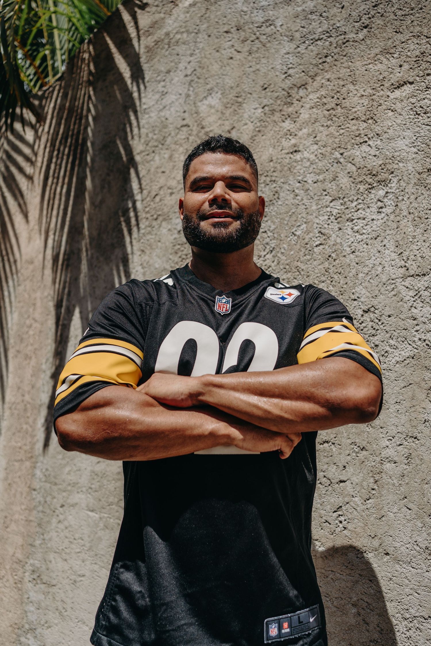 Professional football player in a black NFL Steelers jersey poses with arms crossed against concrete wall.