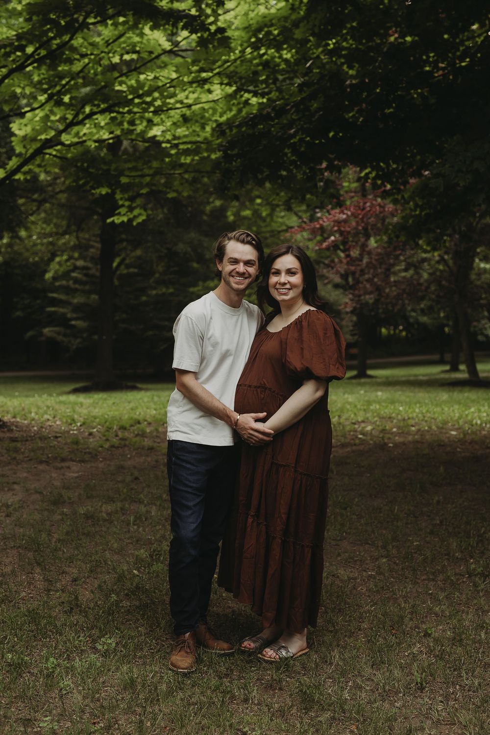 A couple stands together in a wooded outdoor setting wearing casual clothing against a natural backdrop.
