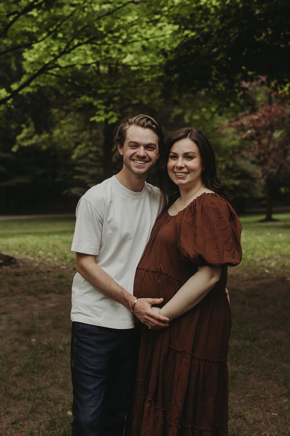 A couple stands together in a forest setting, with the person in brown wearing a flowing tiered dress.