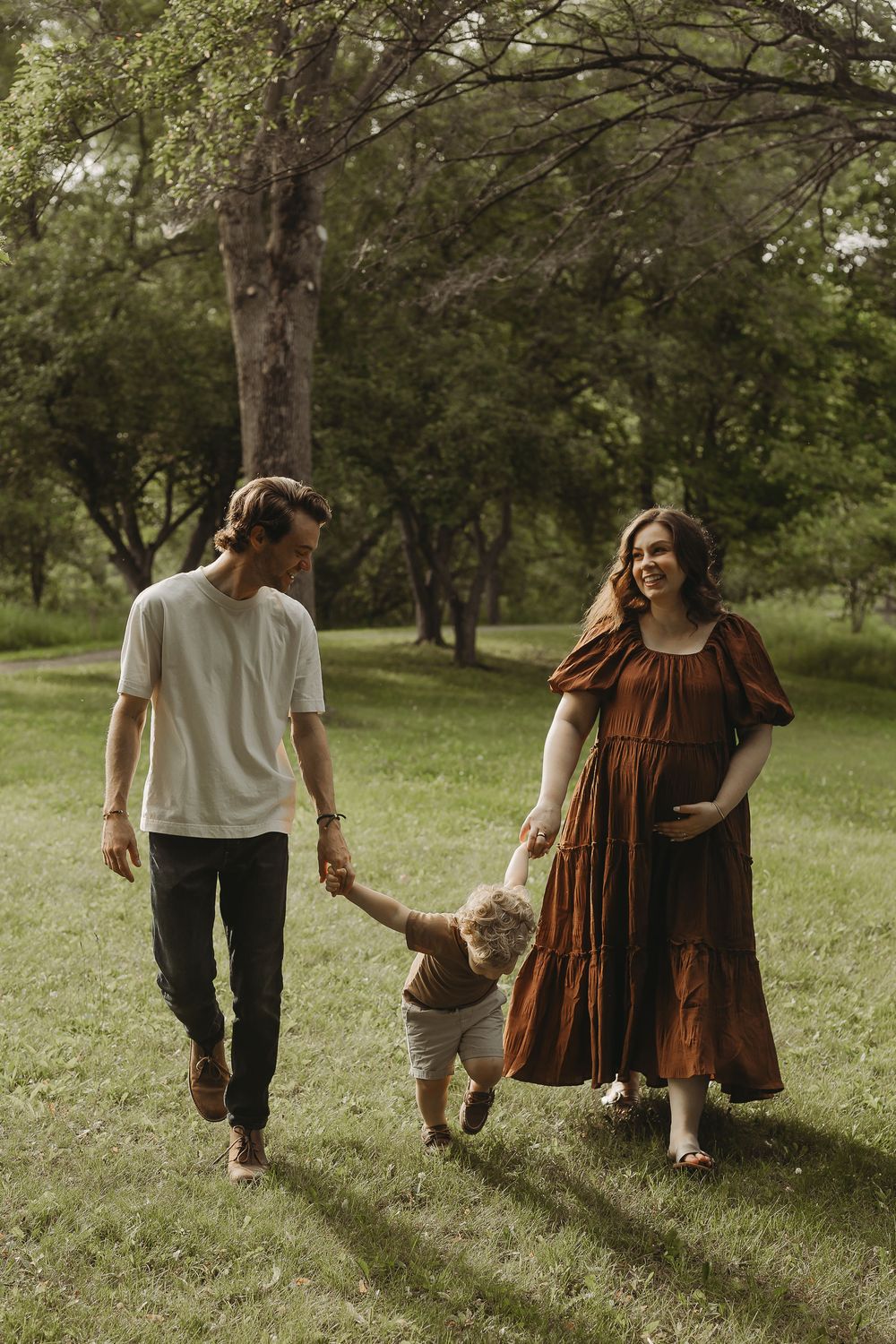 A family strolls through a serene park with tall trees, the mother wearing a flowing rust-colored dress.