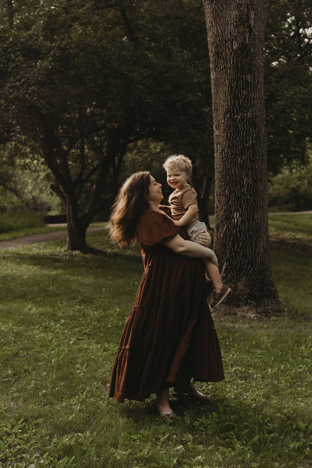Dancing figures in brown dresses twirl together against a backdrop of trees in a sunlit meadow.