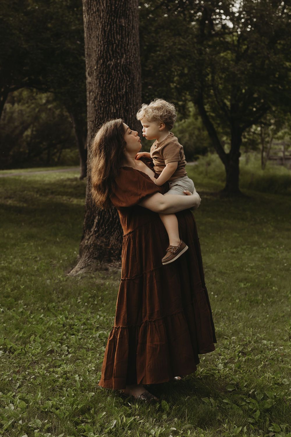 Intimate outdoor portrait of mother and child sharing tender moments under tall trees in a park.