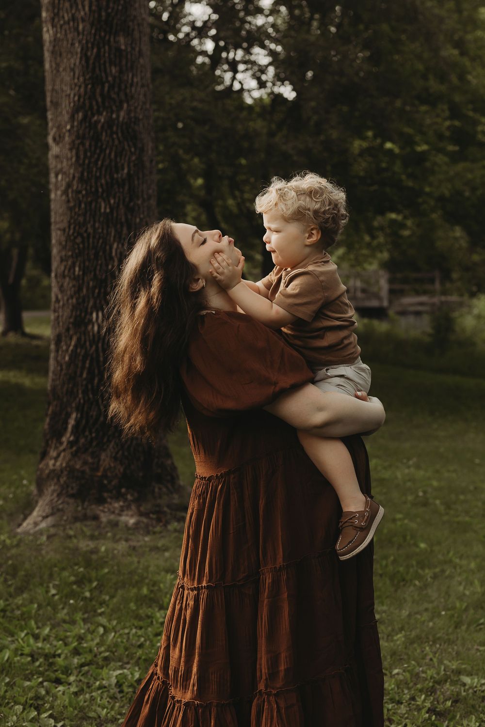 A mother in a brown dress spins and plays joyfully with her child in a green park setting.
