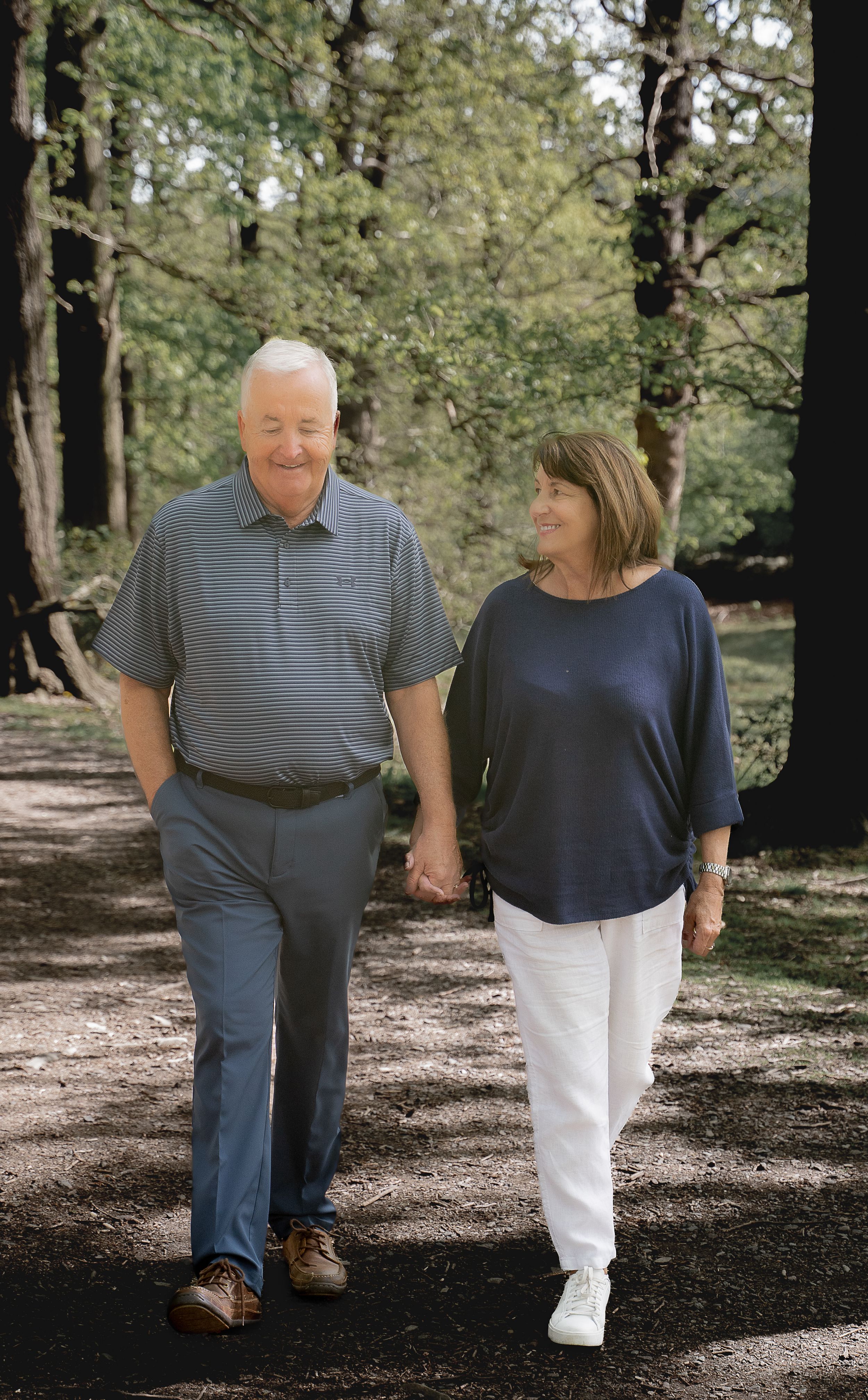 A couple in casual attire walking together through a wooded park on a sunny afternoon.