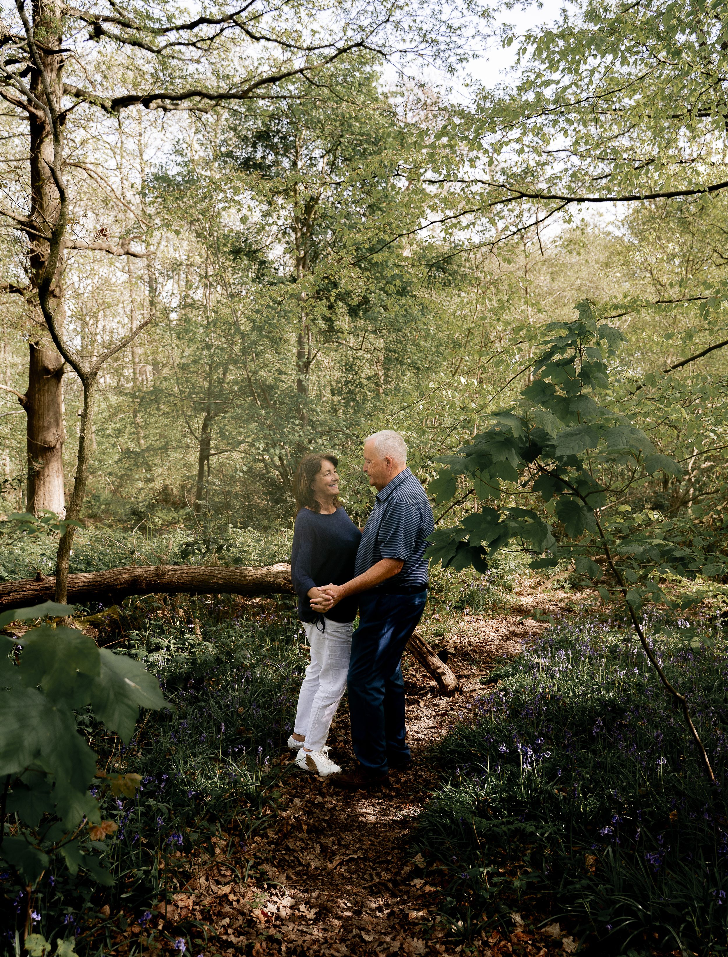 A couple shares an intimate moment while standing on a wooded forest trail surrounded by lush greenery.