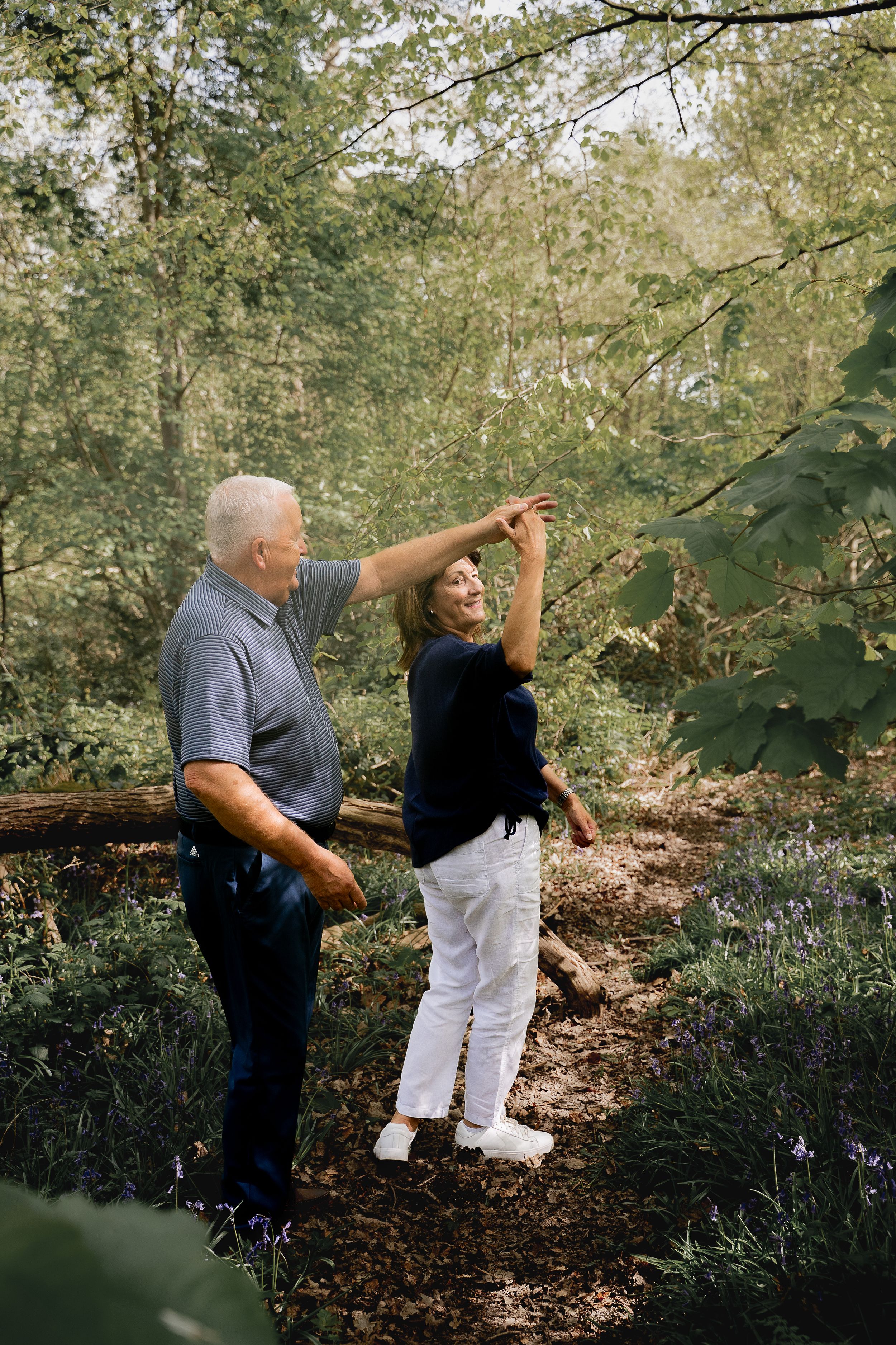 Two people walking together on a wooded path surrounded by lush green foliage and fallen leaves.