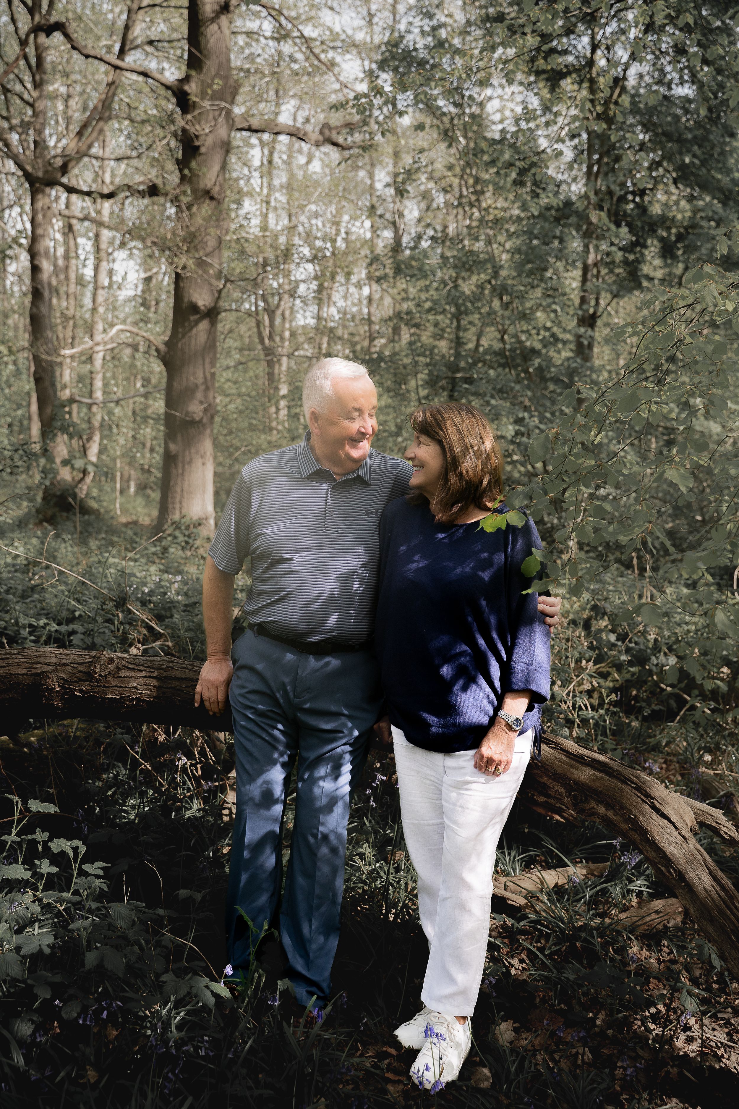 A couple embraces while sitting together on a fallen tree trunk in a wooded forest setting.