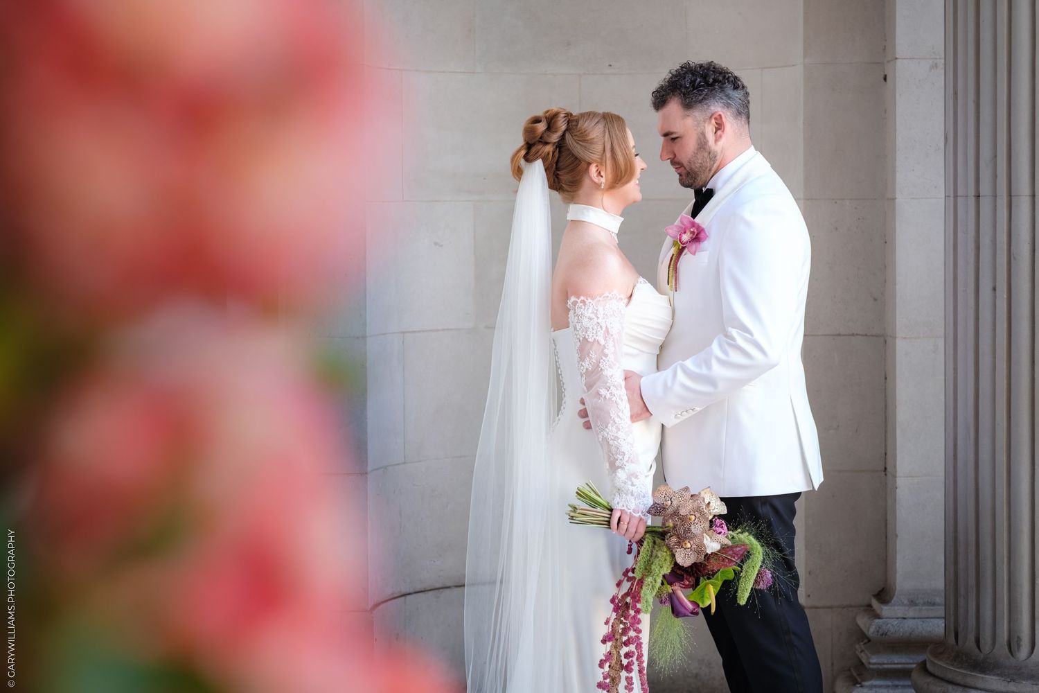 A wedding couple shares an intimate moment together in a marble-columned hallway with soft pink floral details.