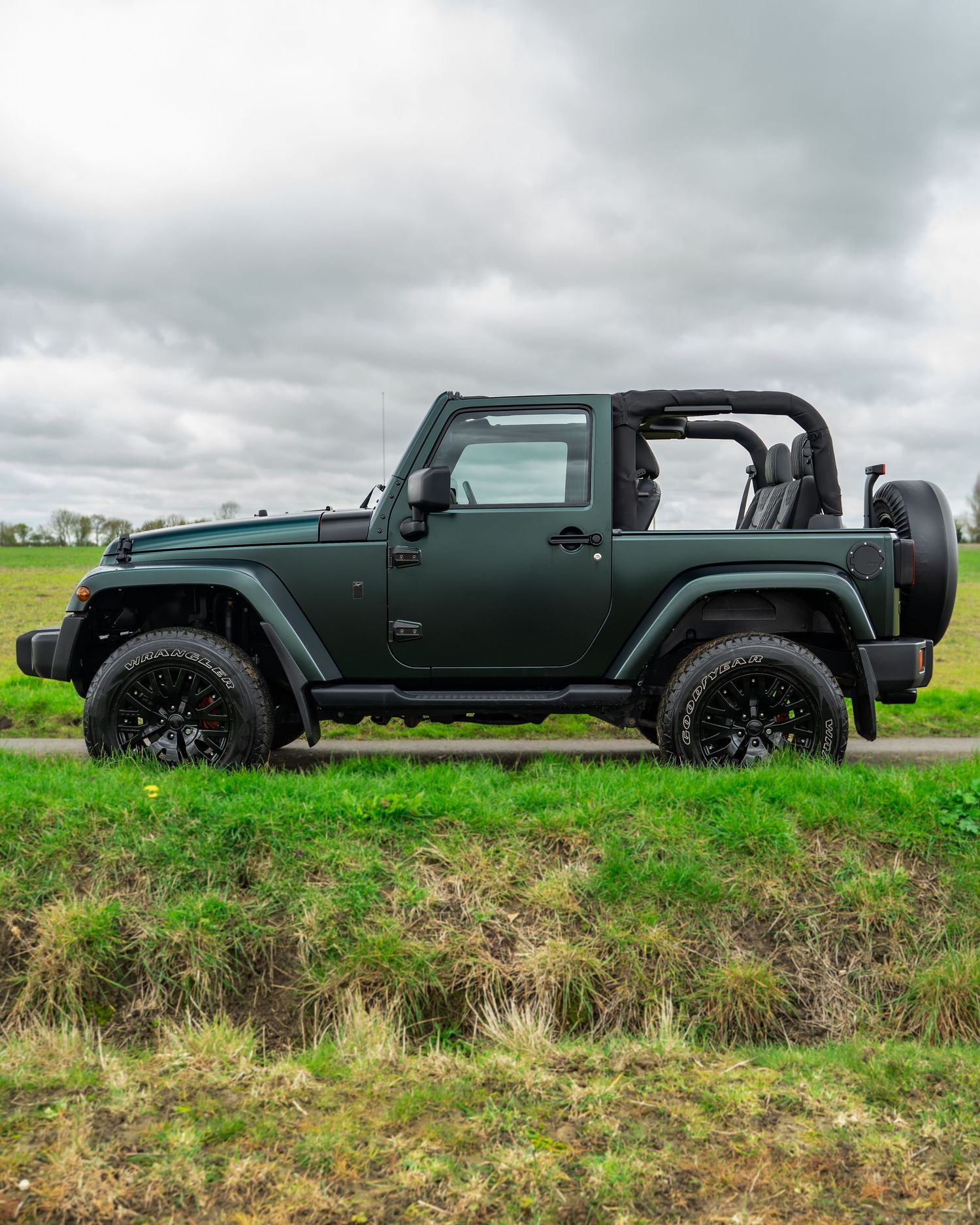 Three black Jeep Wranglers lined up on a grassy hill against cloudy sky.
