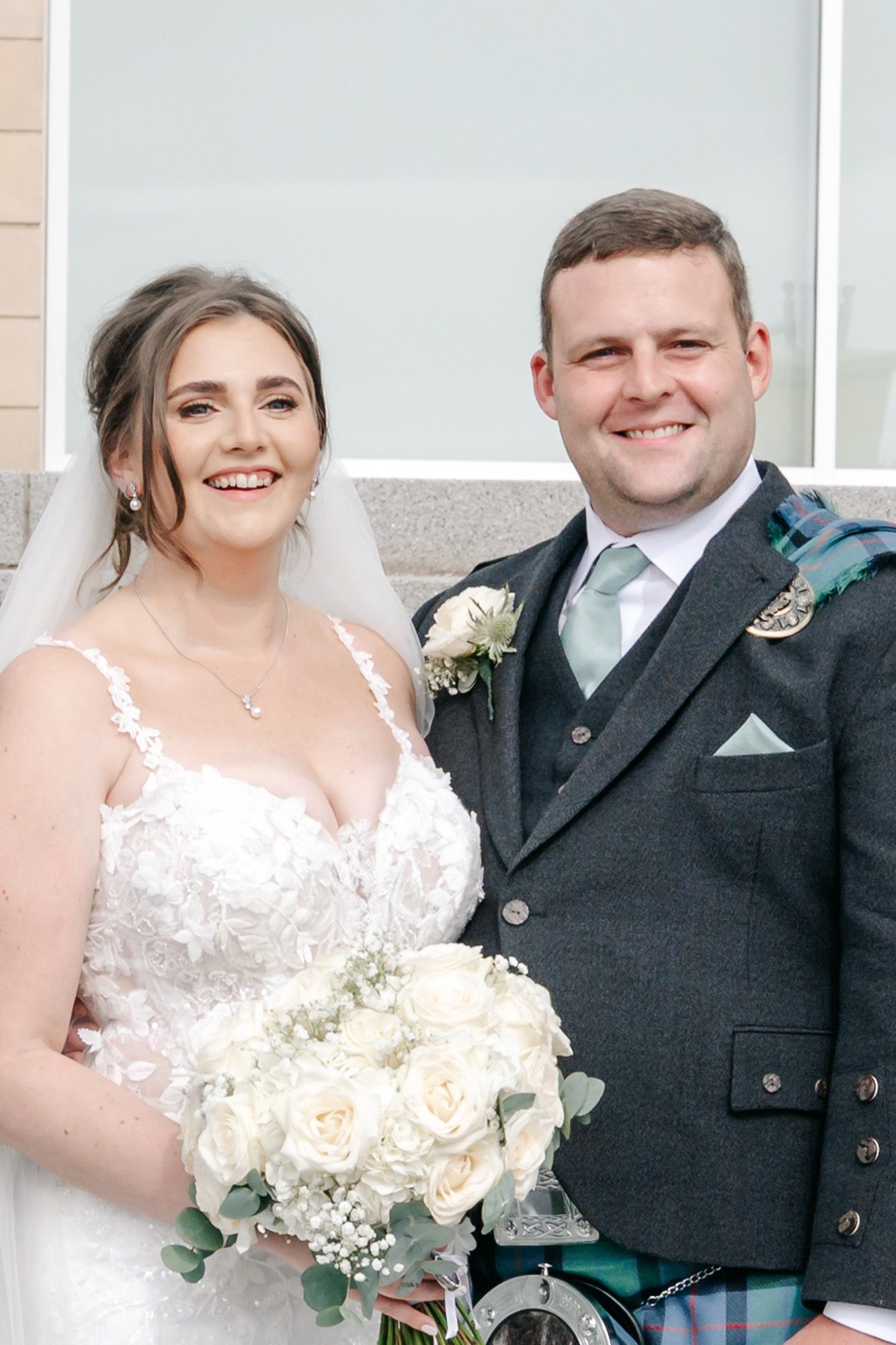 Newlyweds in traditional Scottish and white wedding attire smile together during photo session.