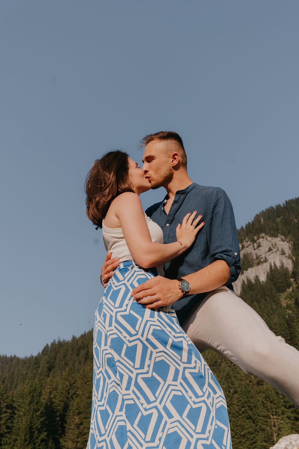A couple shares tender moments against mountain scenery wearing blue and white outfits.