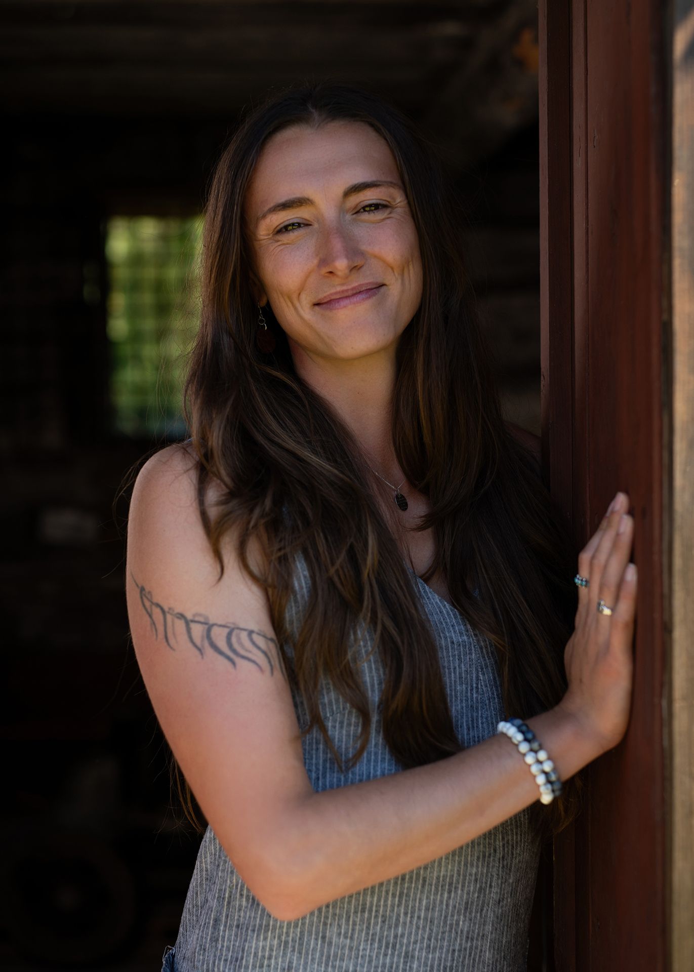 Young woman with long brown hair in pinstripe jumpsuit, tattoo on arm, smiliing in doorway - Okanagan portrait by Tania Veitch.