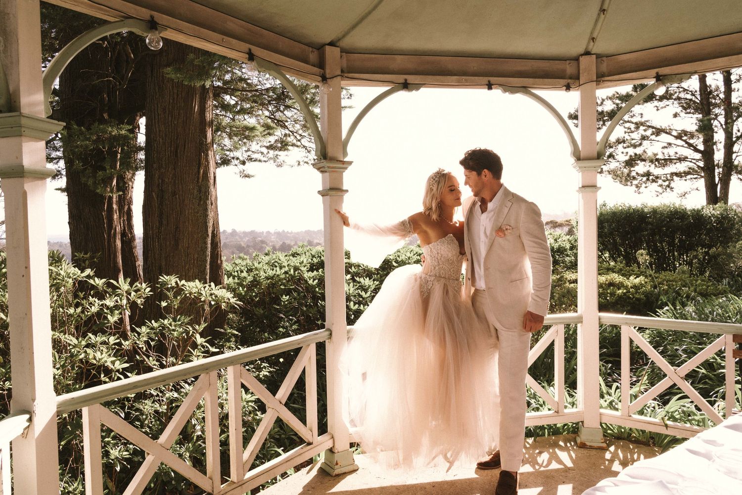 Two people sharing their first dance as newlyweds beneath a white gazebo overlooking scenic countryside views.