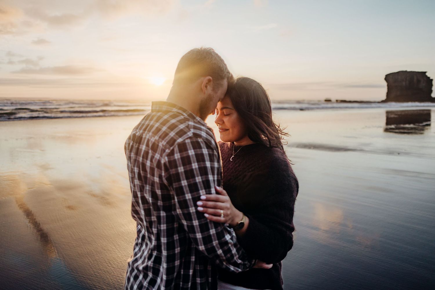 Couple embraces on a serene beach during golden sunset with waves rolling onto shore in background.