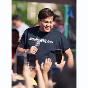 Man in a #BeingFilipino t-shirt smiling and waving to a cheering crowd at a Filipino cultural celebration.