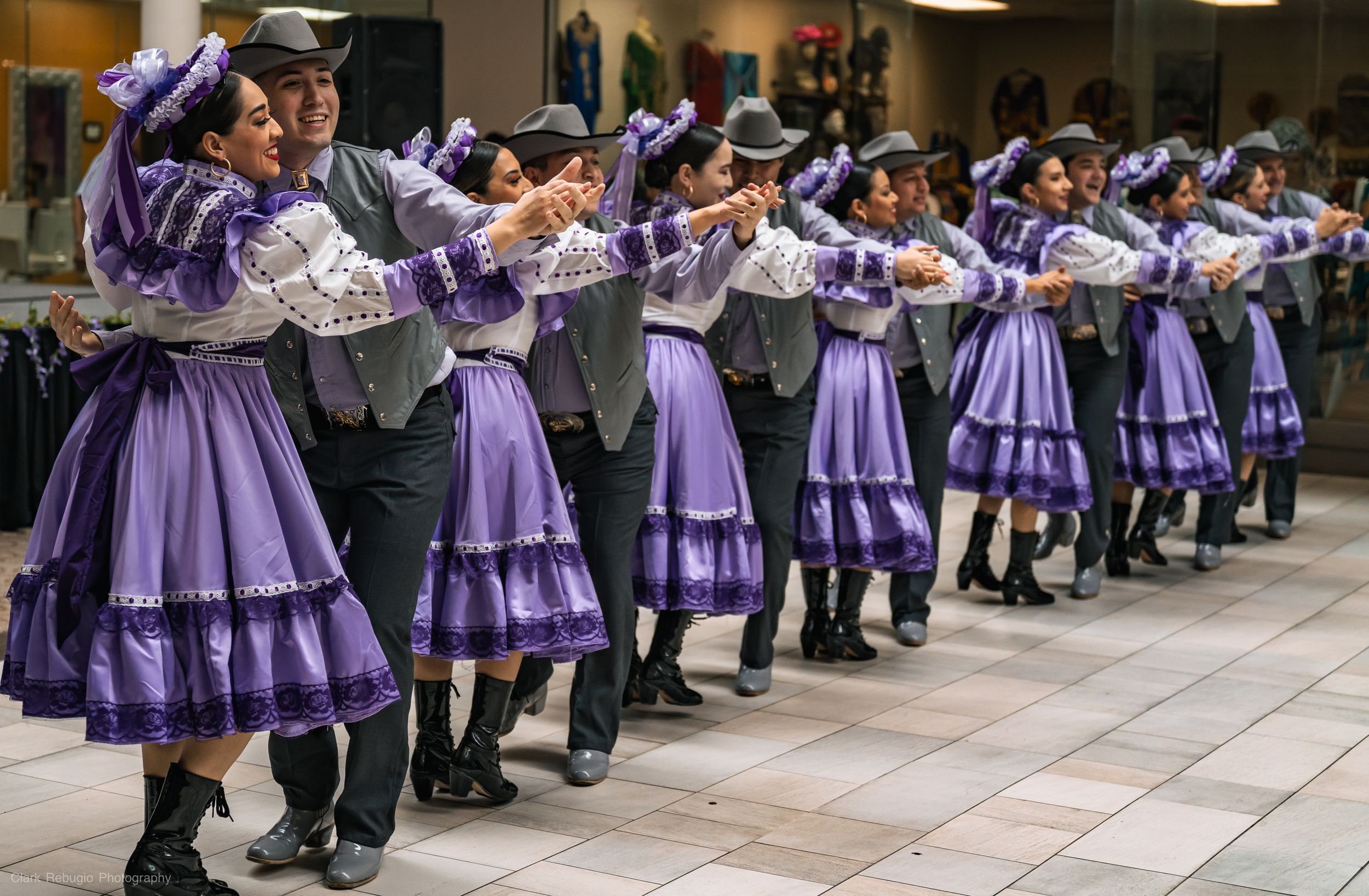 Long line of folkloric dance couples in matching purple and gray costumes performing synchronized steps at a multicultural event.