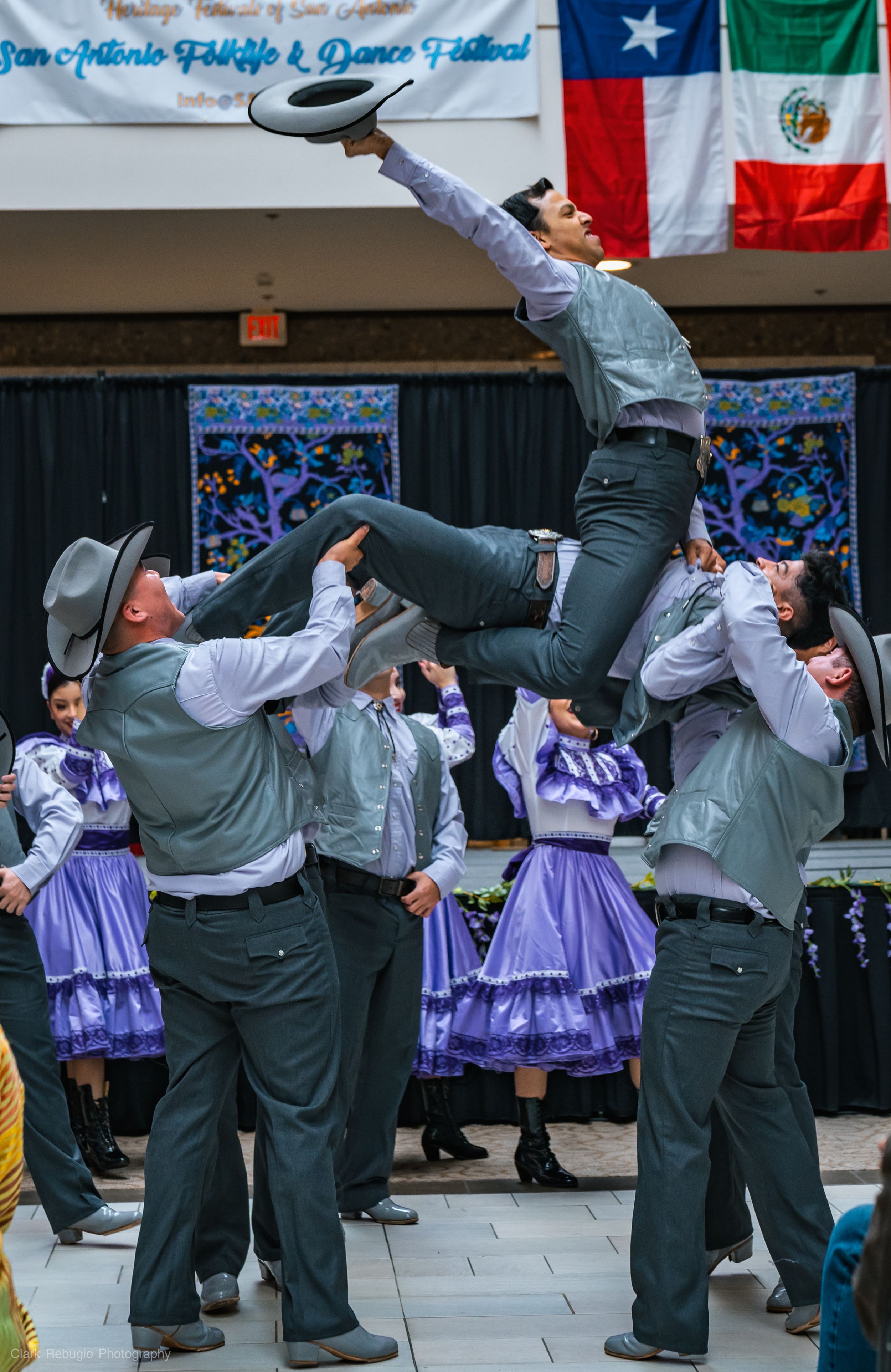 Male folkloric dancers in gray charro outfits lifting their hats while performing a lively traditional dance with women in purple dresses at