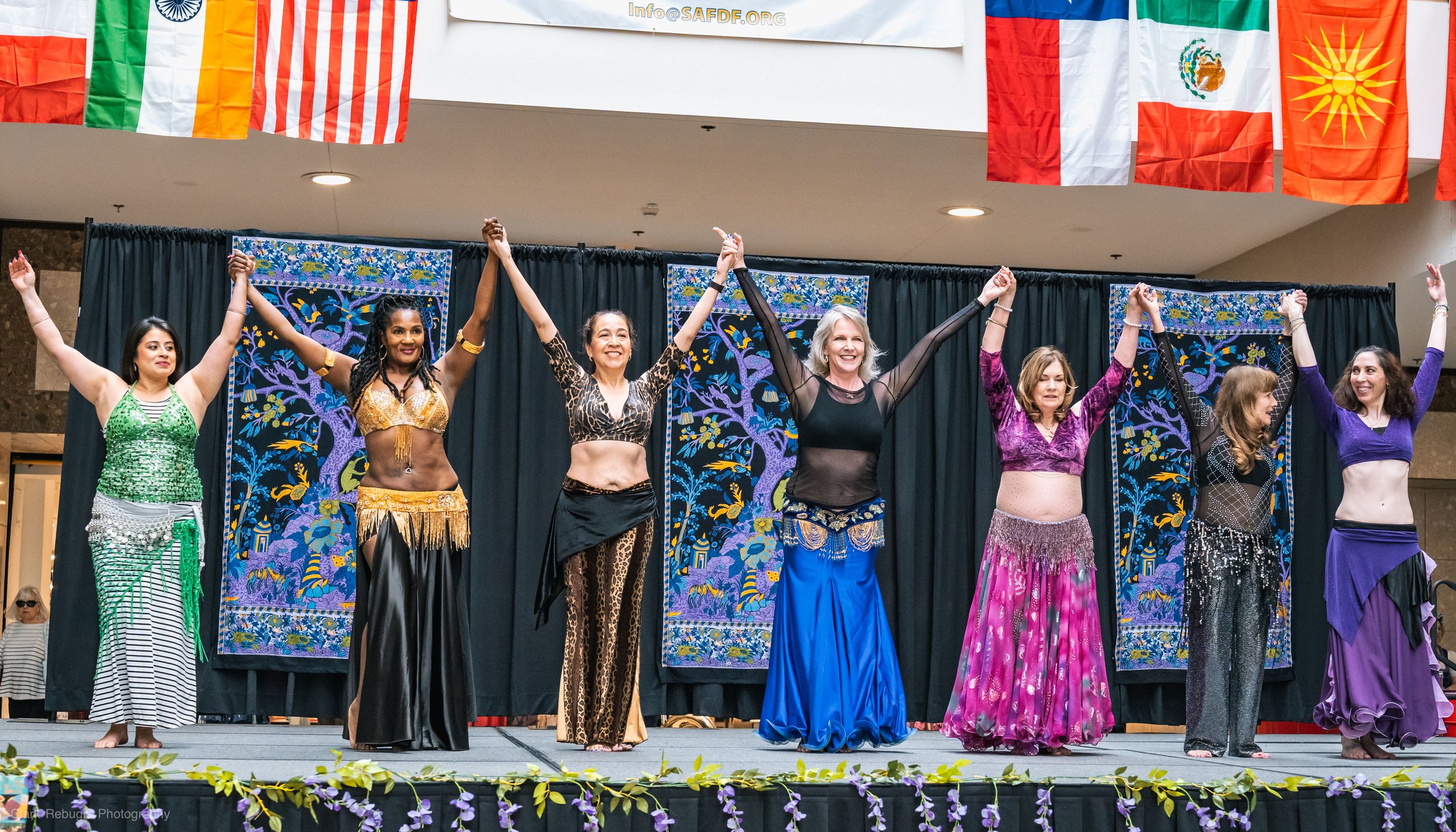Belly dancers in colorful costumes holding hands and taking a bow on stage after a performance at a cultural festival.