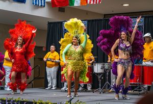 Three samba dancers in red, yellow and purple feathered outfits performing with live drummers at a cultural festival.