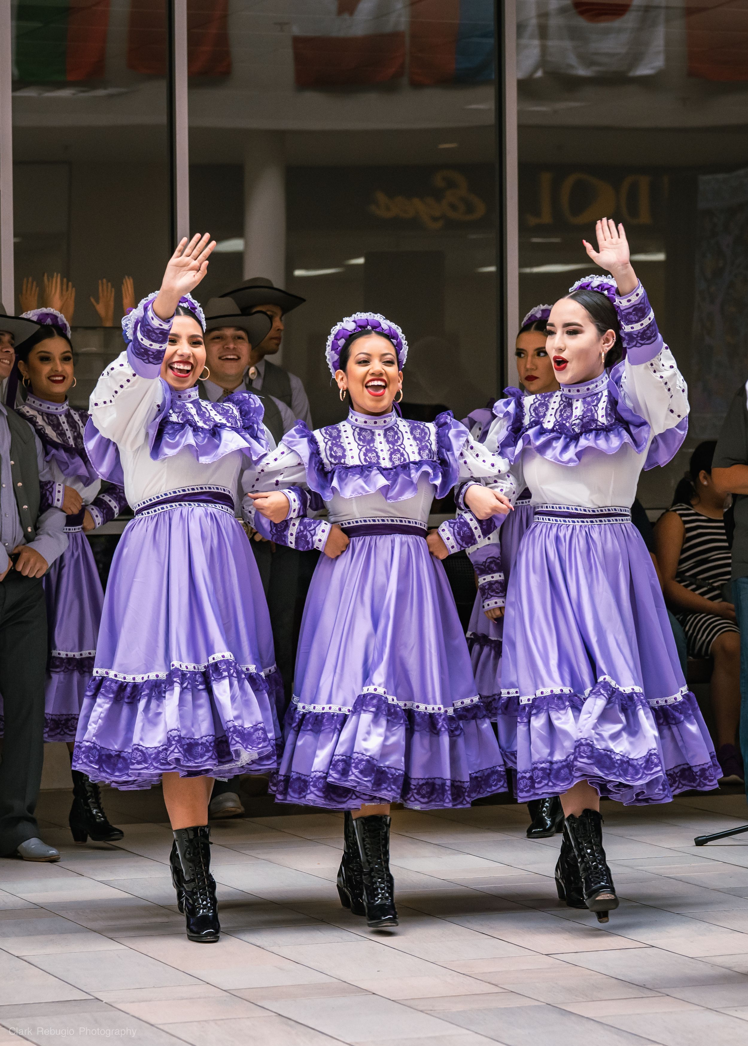 Group of female folkloric dancers wearing matching purple dresses and boots posing together during a multicultural event.
