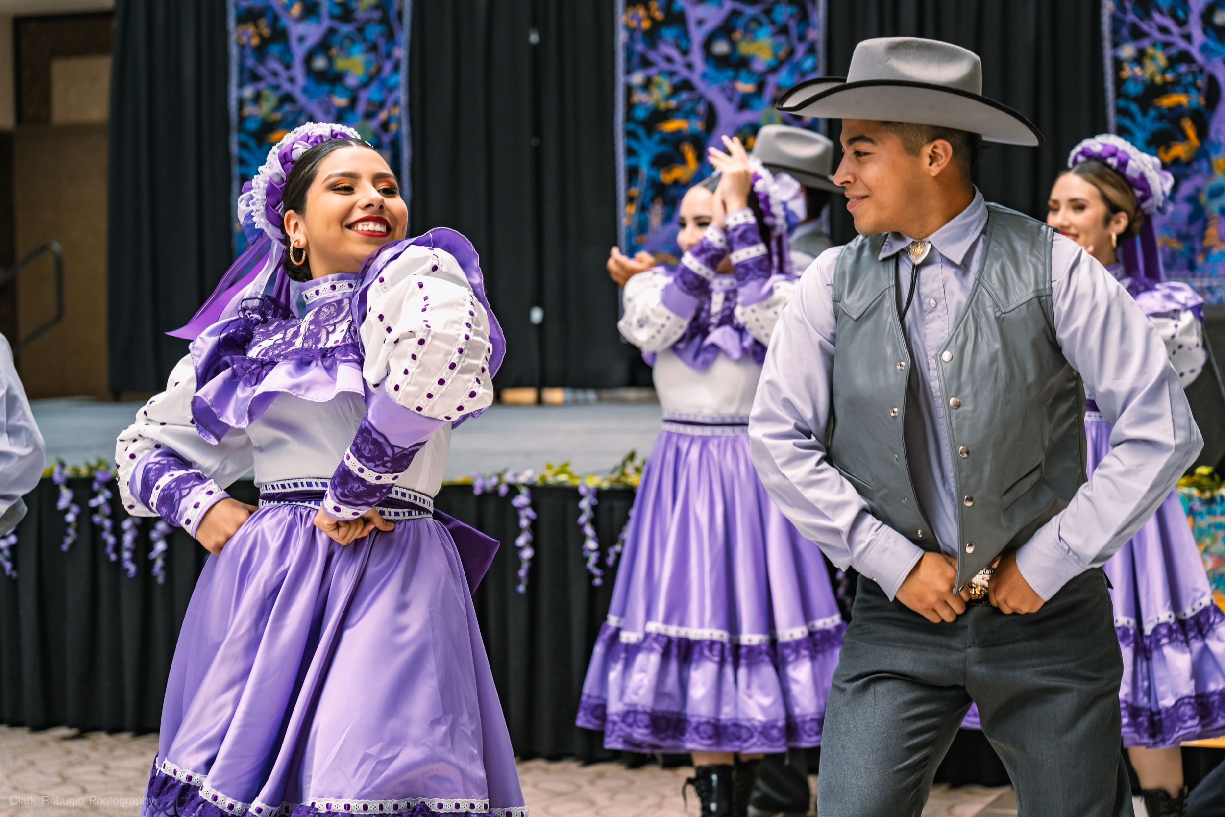Male and female dancers performing a lively folkloric dance in traditional purple costumes on stage at a cultural festival.