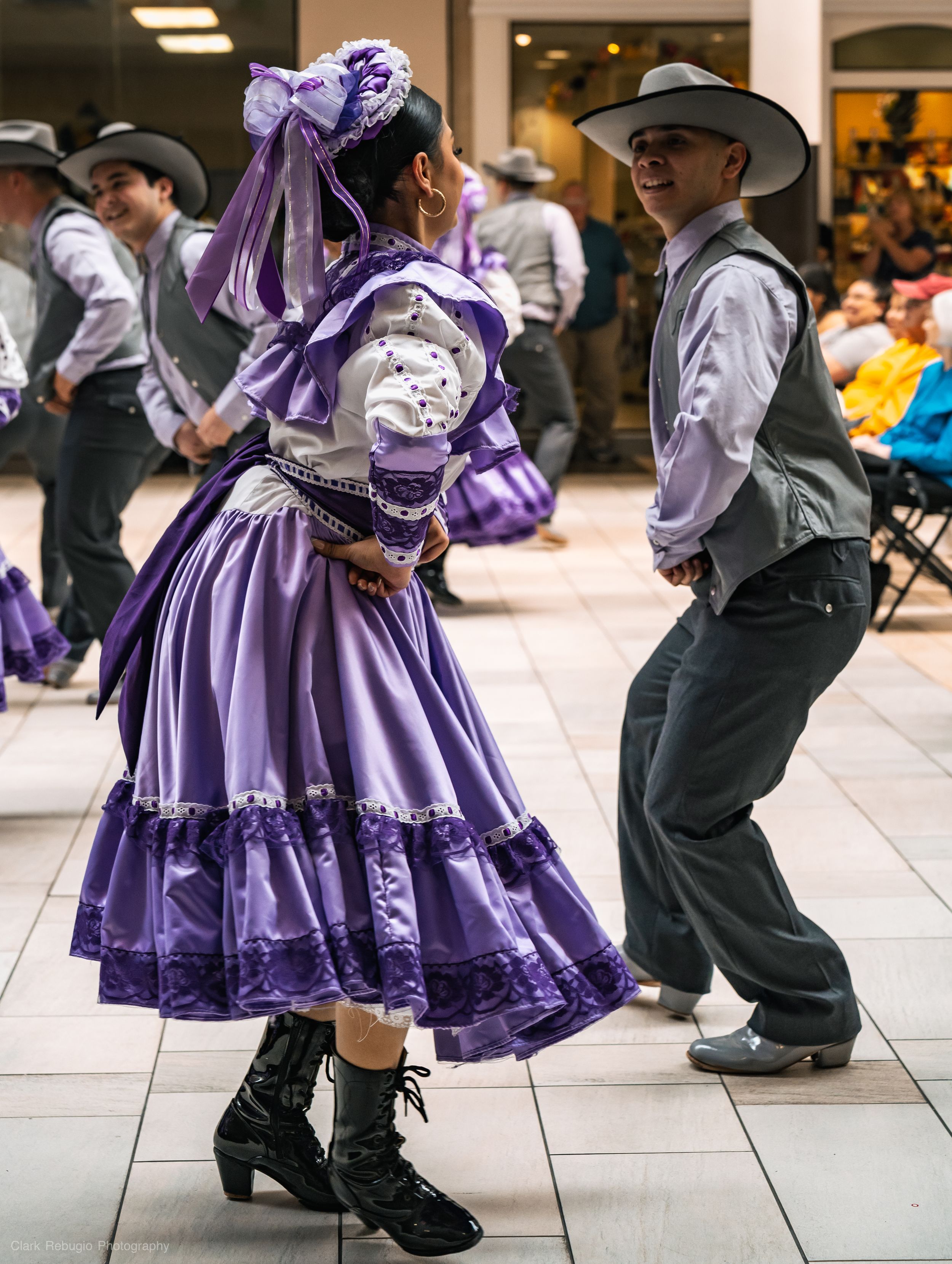 Folkloric dance couple in purple dress and gray charro attire smiling while performing a traditional dance at a cultural festival.