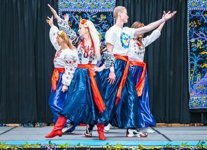 Male and female folk dancers in embroidered white tops and blue skirts with red sashes performing a lively Eastern European style dance.