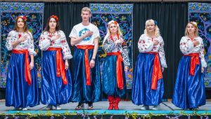 Folk dance troupe wearing traditional Eastern European style costumes with blue skirts, white blouses, and red sashes posing on stage.