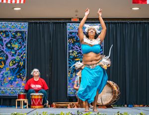 Solo Polynesian dancer in a blue outfit raising her arms while performing a traditional dance with live drumming at a community event.