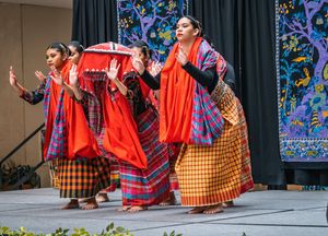 Group of Filipino dancers in red and orange plaid skirts performing a traditional basket dance on stage at a community event.