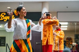 Filipino folk dancers in barong and patterned skirts performing a traditional dance with glasses balanced on their heads.