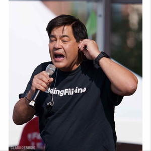 Man in a #BeingFilipino t-shirt speaking passionately into a microphone during an outdoor Filipino community event.