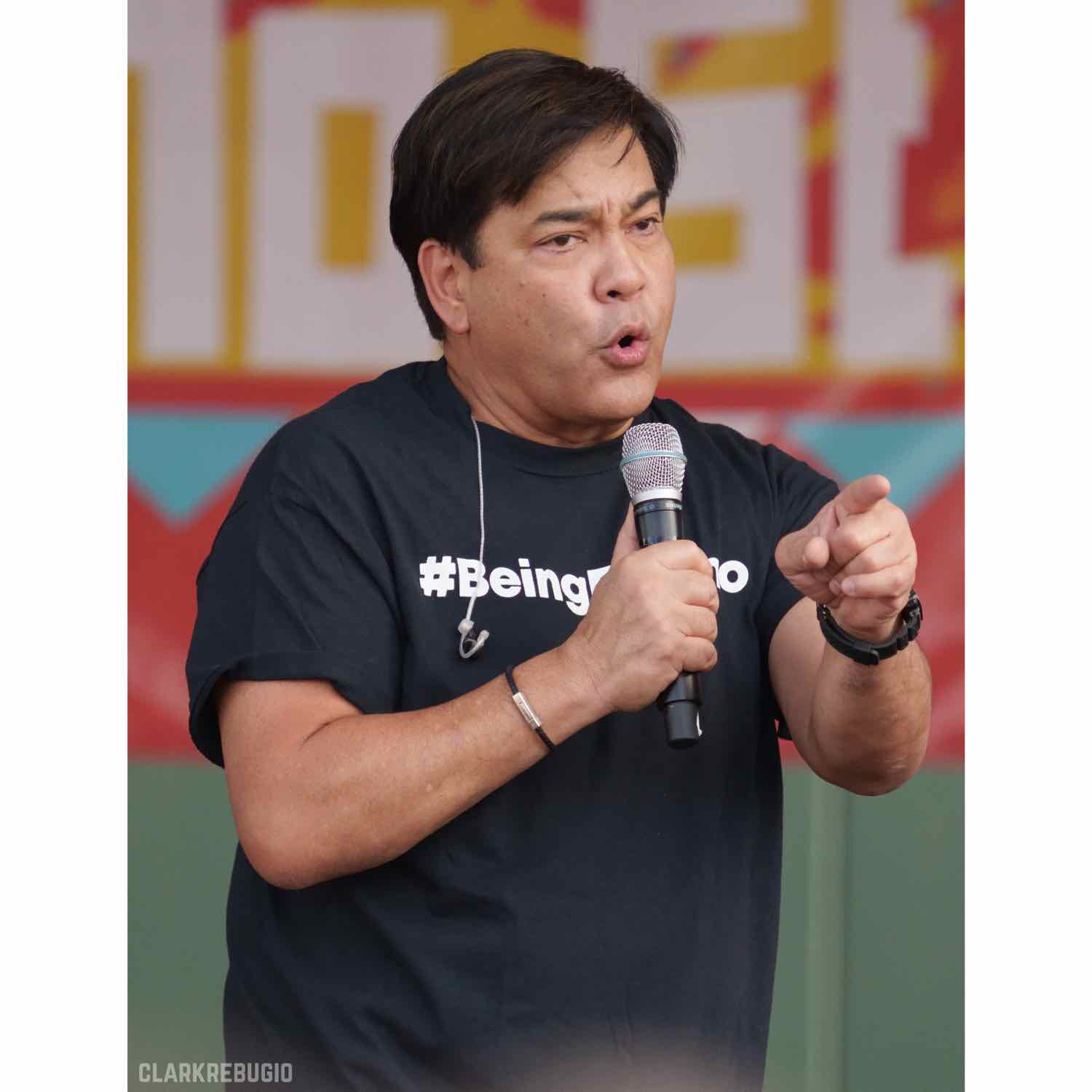 Man in a #BeingFilipino t-shirt speaking to an audience with a microphone at a Filipino cultural event.