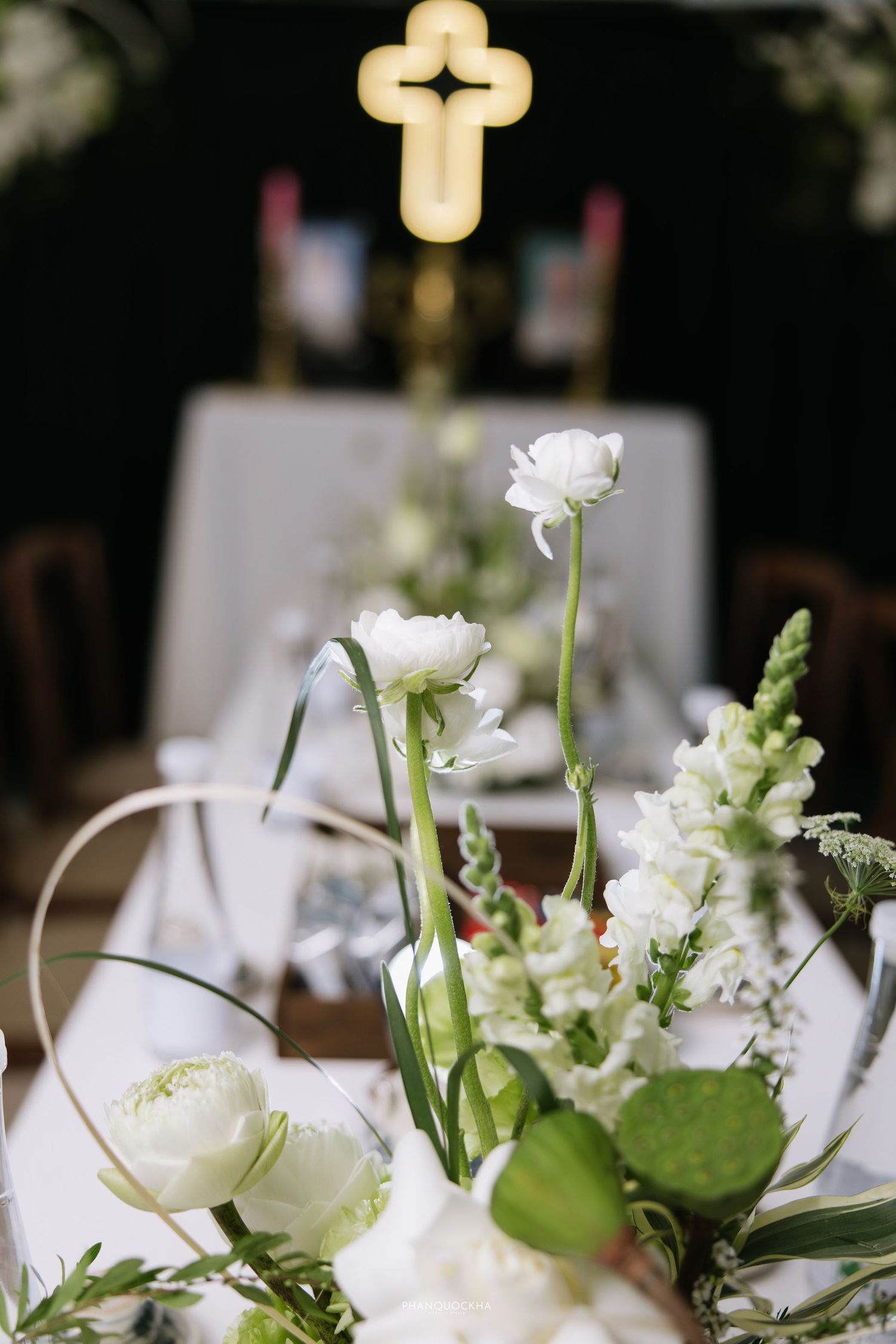 White floral wedding arrangement with roses and snapdragons with illuminated cross on church altar in background.