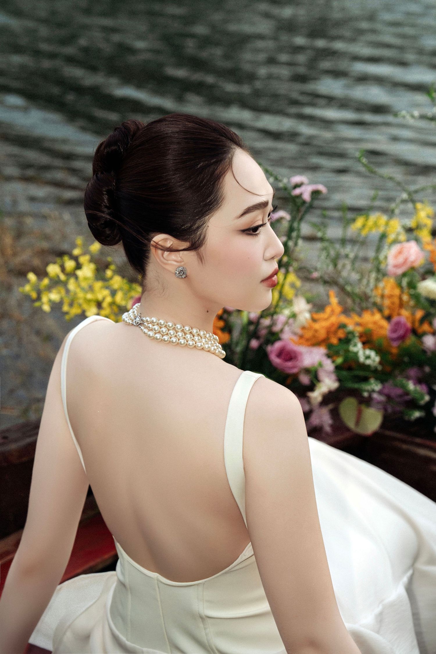 Elegant bride in backless white gown with pearl necklace seated near colorful flowers by a lake.