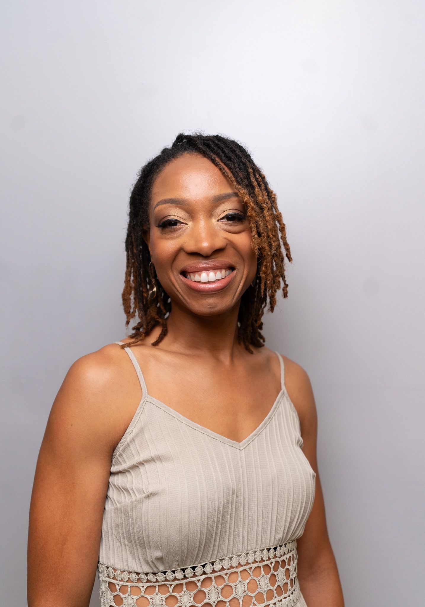 A woman wearing a beige crochet-trimmed tank top smiles warmly against a light gray background.